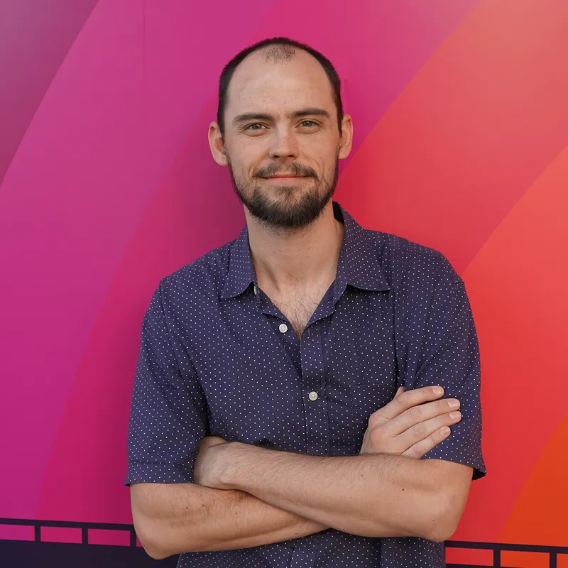 Andrew Riefenstahl smiling with his arms crossed against a multicolored background with hues of purple and maroon to pink.